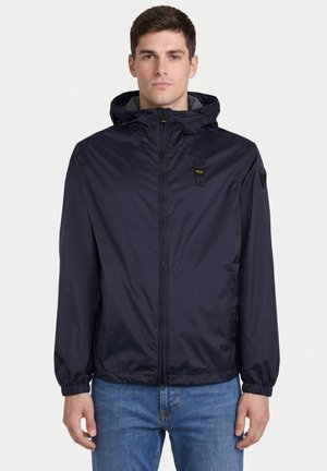 Mężczyzna w granatowej bluzie z kapturem na suwak z elastycznymi mankietami i niebieskich dżinsach, stojący na białym tle.