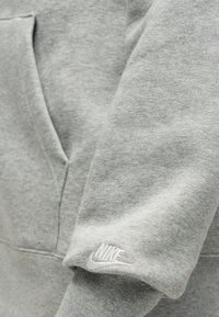 Zbliżenie na szary rękaw bluzy Nike z haftowanym białym logo Nike i przednią kieszenią kangurzą.