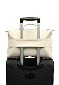 Kremo spalvos odinė rankinė su viršutinėmis rankenomis, padėta ant juodo lagaminų su ratukais. Rankinė pritvirtinta prie lagamino išsikiančios rankenos.