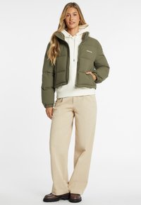 Žalias pūkuotas švarkas, trumpas dizainas, užtrauktukas priekyje, siūtas tekstūra. Dėvimas ant baltos džemperio su gobtuvu ir derinamas su kreminiais plačiais kelnais.