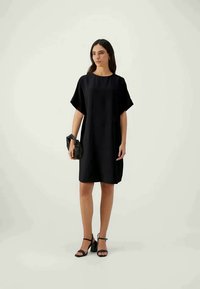Vestido preto de corte simples com mangas curtas, ajuste ligeiramente solto, textura suave e decote redondo. Combinado com uma carteira preta e saltos.