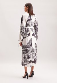 Langes weißes Kleid mit schwarzem architektonischen Druck, das über lange Ärmel, eine taillierte Taille und Bindebänder an den Ärmeln verfügt. Kombiniert mit schwarzen Absätzen.