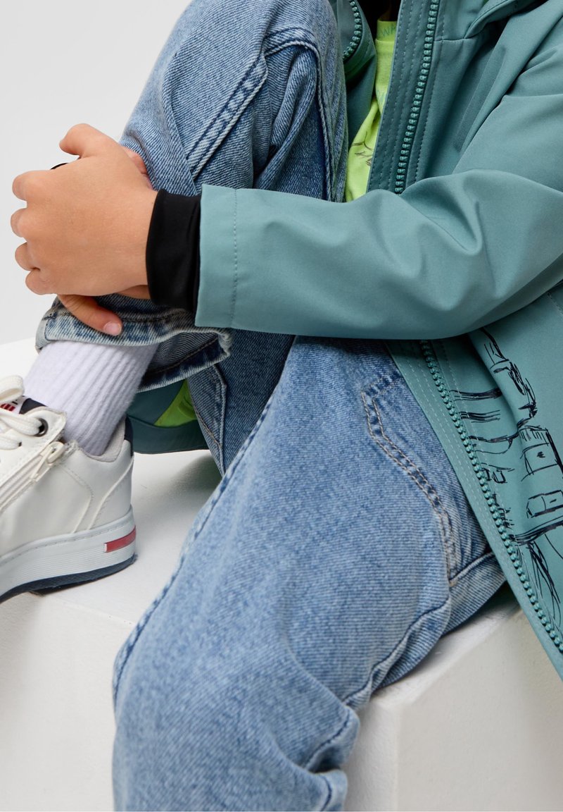 Enfant portant des baskets blanches, des chaussettes blanches, un jean bleu et une veste bleu sarcelle, assis avec les genoux ramenés vers le haut et les bras entourant les jambes.