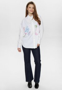 Cămașă albă cu nasturi, decorată cu broderie florală colorată în roz, albastru și verde; asortată cu blugi negri evazați și cizme cu toc negre.