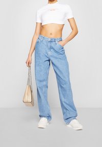 Kvinna som bär ljusblå vida jeans, vit kortärmad T-shirt med logotyp, vita sneakers, och håller en beige handväska med silverkedja.