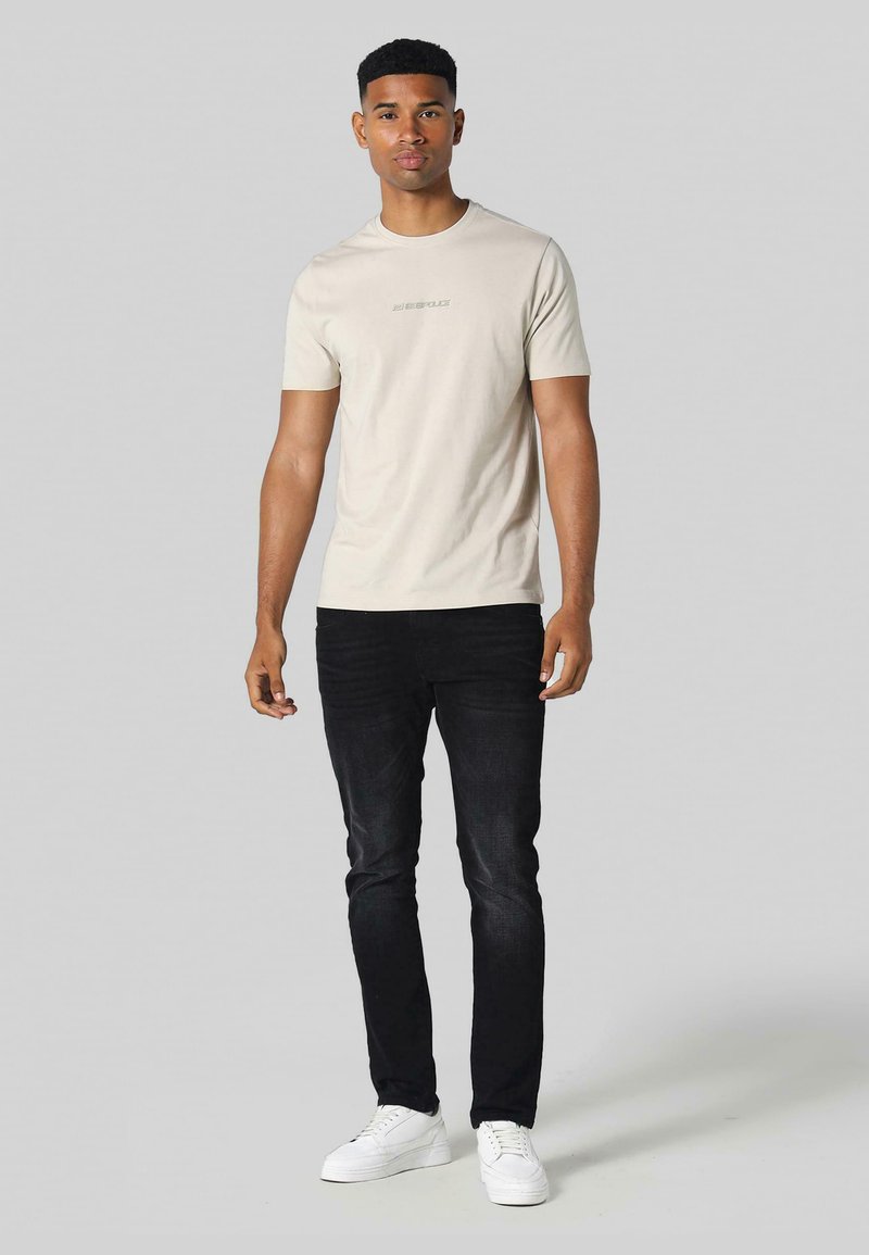 Béžové tričko s krátkými rukávy a kulatým výstřihem, které má na hrudi diskrétní logo. Spárované s černými slim-fit džínami a bílými teniskami.