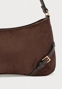 Καφέ σουέντ τσάντα χειρός με ομαλή υφή, καμπυλωτό σχήμα και δερμάτινο λουράκι με διακοσμητικό αγκράφα σε χρυσή απόχρωση.