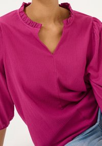 Blouse magenta avec encolure à volants, manches courtes ballon et découpe en V. Tissu texturé avec des rayures verticales.