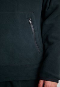 Tamsiai žalias fliso puloveris su užtrauktuku kairėje pusėje. Minkšta tekstūra ir apvadas, pasižymintis paprastu, funkcionaliu dizainu.