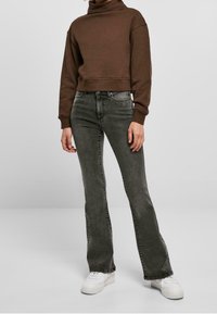 Persona con sudadera marrón corta, jeans acampanados de talle alto y negro descolorido, y zapatillas blancas sobre un fondo gris liso.