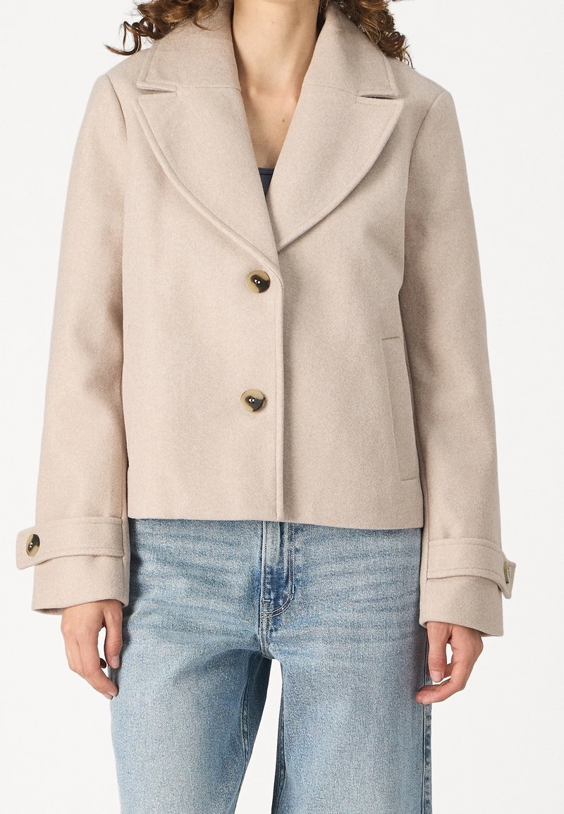 Manteau court en laine beige avec larges revers et deux boutons, porté sur un jean bleu clair.