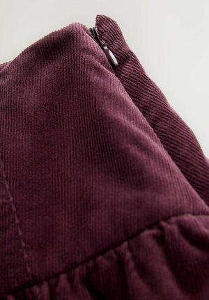 Lähiotoksella tumma violetti corduroy-kangas, jossa näkyy uurteet ja osittain vedetty vetoketju valkoista taustaa vasten.