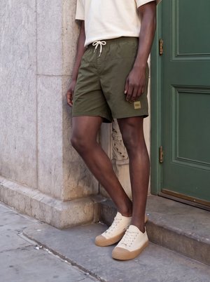 Homme portant un short vert olive, des chaussures beige décontractées et une chemise de couleur claire, debout près d’un mur en pierre et d’une porte verte.
