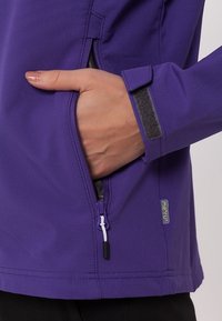 Lila Softshell-Jacke mit einer Reißverschlusstasche und einem Klettverschluss am Ärmel. Angenehme Textur mit einem elastischen Bund am Saum und gesticktem Logo.