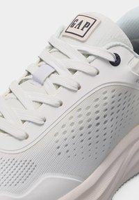 Κοντινή λήψη λευκού αθλητικού παπουτσιού GAP με δίχτυ στις πλαϊνές επιφάνειες, λευκά κορδόνια και ετικέτα με το λογότυπο GAP στη γλώσσα του παπουτσιού.