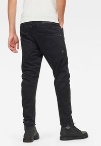 Pantalones de mezclilla negra con un corte ajustado, que presentan bolsillos traseros en ángulo, una textura sutil y costuras reforzadas. Combinados con botas negras con cordones.