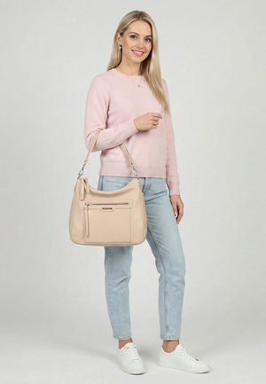 Vrouw in een roze trui, lichtblauwe jeans, witte sneakers, glimlachend en een beige handtas met ritssluiting over de arm houdend op een grijze achtergrond.