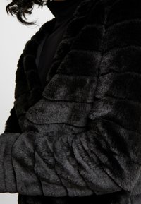 Black faux fur jacket with a textured wavy pattern, featuring a shawl collar and long sleeves. Soft, plush surface with a draped design.
