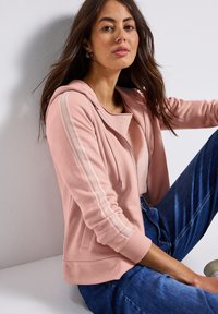 Hoodie rosa chiaro con zip e cappuccio, caratterizzato da strisce bianche perforate sulle maniche. Abbinato a jeans di jeans blu. Texture liscia.