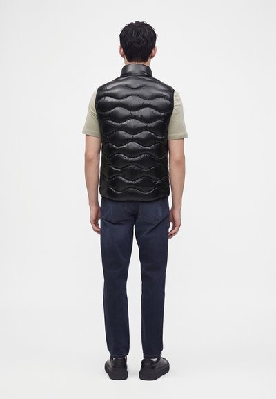 Hombre con chaleco negro acolchado sobre camisa clara de manga corta, vaqueros oscuros y zapatillas negras, de pie de espaldas sobre fondo blanco.
