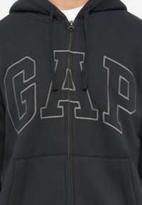 Person som bär en svart zip-up-hoodie med stort grått 'GAP'-logotyp, dragsko och en vit t-shirt under.