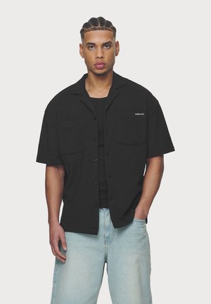 Μαύρο κοντομάνικο πουκάμισο με υφασμάτινη υφή, δύο τσέπες στο στήθος, κουμπί στο μπροστινό μέρος, σε συνδυασμό με ανοιχτόχρωμες τζιν παντελόνες.