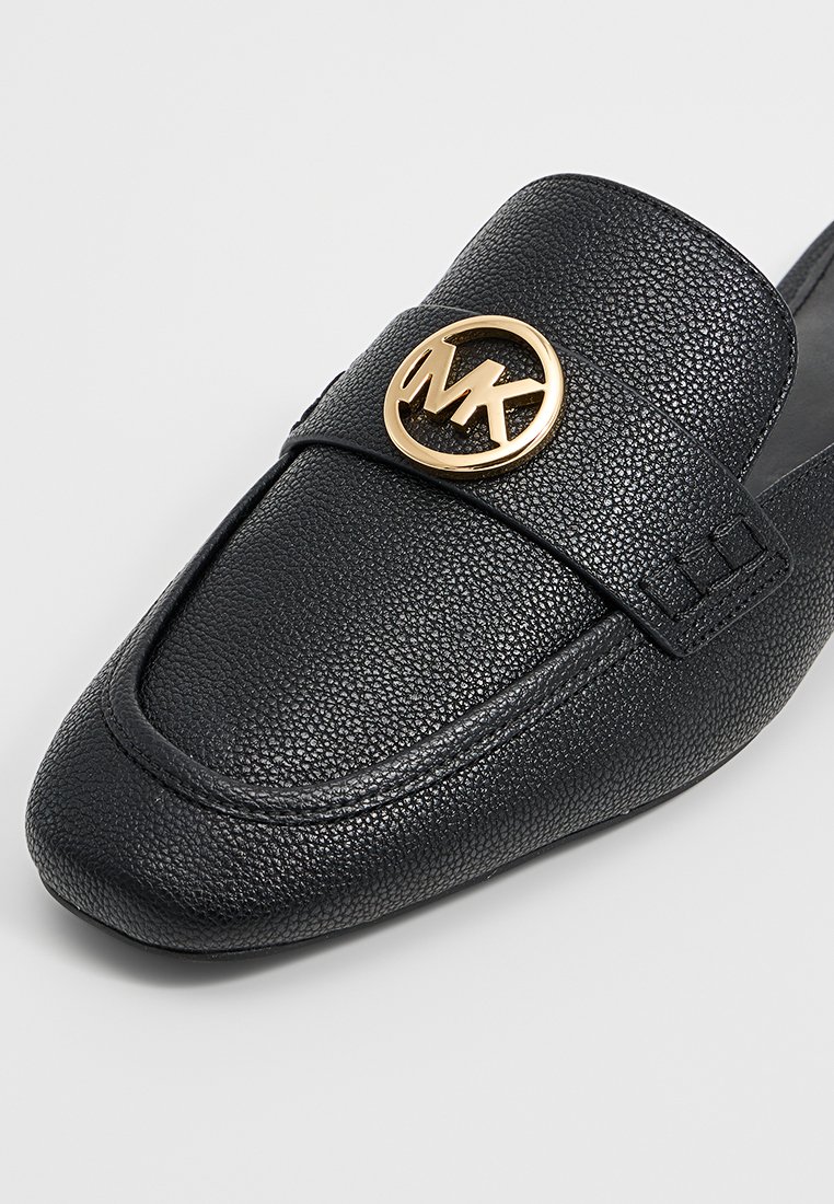 Zwarte leren instapper met een zachte textuur, ronde neus en een goudkleurig cirkelvormig logoaccent op de bovenkant. Strakke, minimalistische uitstraling.