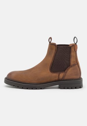 Bota Chelsea de cuero marrón con panel elástico negro en el costado, tiradores y suela de goma negra resistente, mostrada de perfil mirando hacia la izquierda.