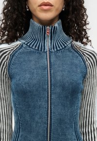 Μπλε πουλόβερ με φερμουάρ και ψηλό λαιμό, με υφή πλέξης και ριγέ μανίκια σε αντίθετες σκούρες και ανοιχτές αποχρώσεις. Λεπτομέρεια ασημένιου φερμουάρ.