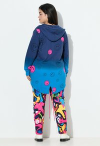 Sudadera con capucha en azul ombré con patrones de caras sonrientes rosas, combinada con pantalones de patrón abstracto colorido que presentan varias formas y colores.