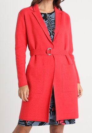 Kobieta w czerwonym, sięgającym kolan płaszczu z paskiem, nałożonym na wzorzystą czarno-niebiesko-czerwoną sukienkę, stojąca z rękami swobodnie opartymi po bokach.