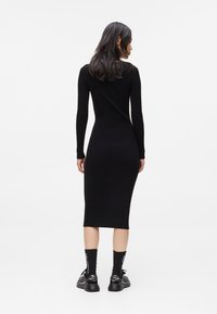 Vestido de malha canelada preto com mangas longas, corpo justo e comprimento midi. Modelo usa sapatos e meias pretas. Design simples e elegante.