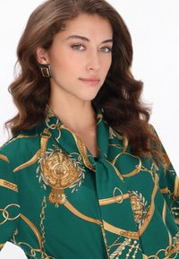 Bluza verde cu un model de lanț auriu, cu guler legat și textură netedă, asemănătoare satenului. Modelul poartă cercei pătrați.