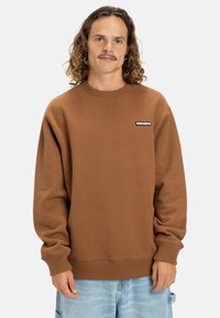 Sudadera marrón con puños y dobladillo acanalados, que presenta un pequeño parche negro de logo en el pecho. Diseño de cuello redondo con una textura suave.