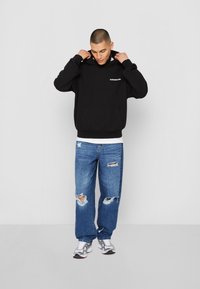 Sweat à capuche noir avec poche avant, associé à un jean en denim bleu usé. Le modèle ajuste la capuche. Les baskets sont argentées.