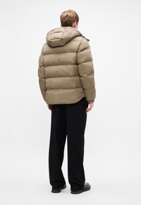 Puffjakke i olivengrøn, med quiltet design, hætte og elastiske manchetter, parret med sorte vide bukser og sorte sko.