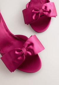 Fuksianväriset satiiniset liukusandaalit, joissa on suuri rusetti edessä, sileä pinta ja litteä pohja.