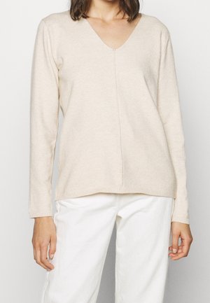 Beige langærmet v-hals sweater lavet af blødt strikket materiale, med en central søm og ribbede manchetter, parret med hvide bukser.