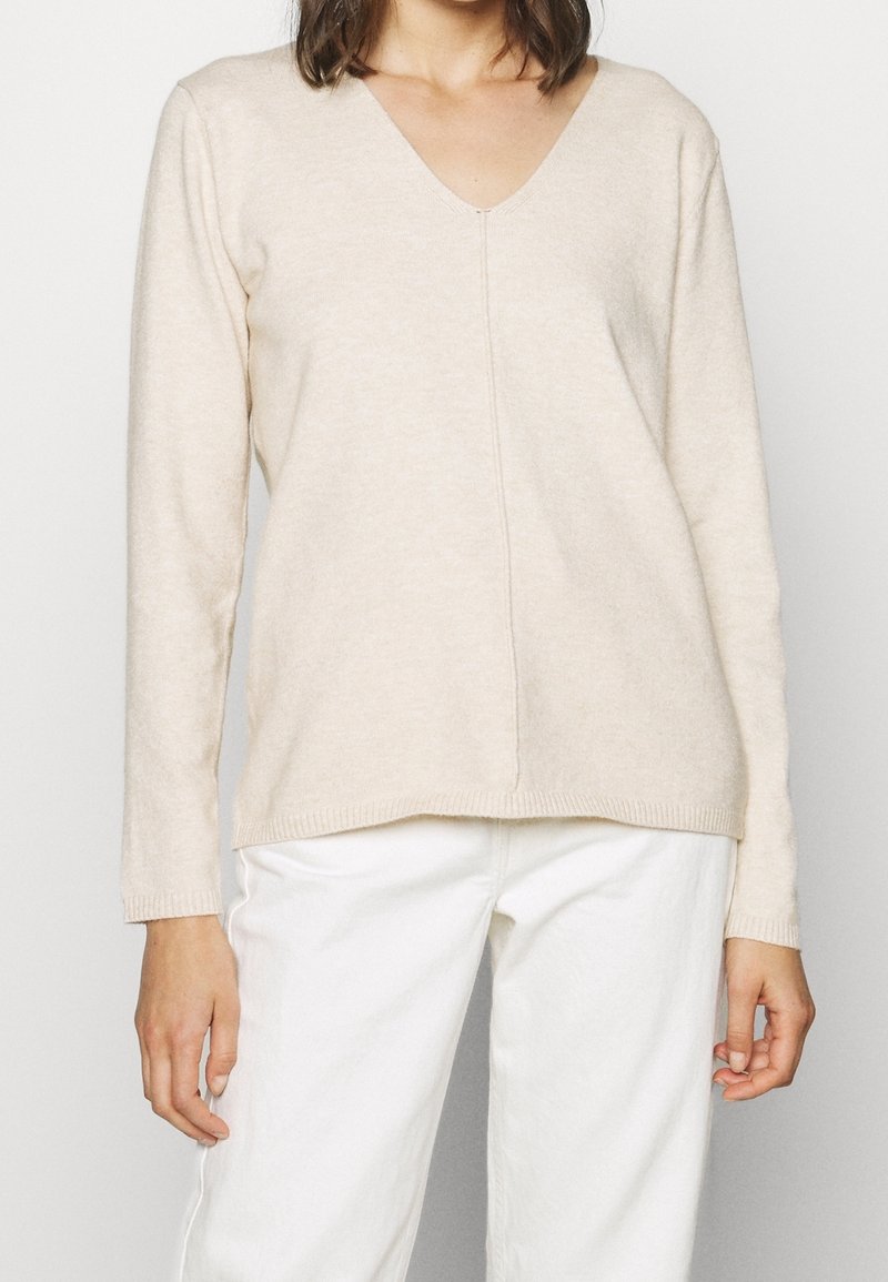Maglione beige a maniche lunghe con scollatura a V realizzato in morbido tessuto a maglia, con una cucitura centrale e polsini a costine, abbinato a pantaloni bianchi.