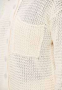 Πλεκτή ζακέτα σε εκρού χρώμα με ανοιχτό υφαντό σχέδιο, που διαθέτει τετράγωνη τσέπη και κούμπωμα με κουμπιά. Μαλακό ύφασμα και αναπνεύσιμος σχεδιασμός.