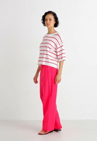 White striped shirt with red lines, relaxed fit. Paired with bright pink, loose-fitting trousers. Model standing on a neutral background.