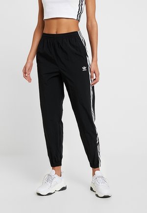 Femme portant un pantalon de survêtement Adidas noir avec des bandes blanches et un logo, associé à des baskets blanches épaisses et un top court blanc.