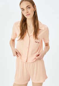 Persistan i persikofärg, ribbad pyjamas-set i kort ärm med en knäppning i toppen som har krage och broderad text "GOOD LUCK", samt matchande shorts.