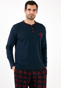 Camisa de manga larga azul marino con un collar de cuadros y botones rojos. Combinada con pantalones de cuadros con líneas rojas y negras. Pequeño emblema rojo en la camisa.