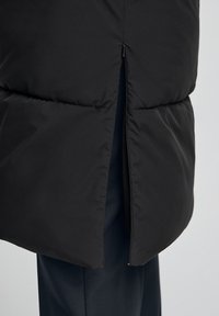 Svart vadderad jacka med en delvis uppdragen sidospalt som visar mörka byxor under.