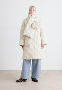 Casaco acolchoado creme com um padrão de diamante acolchoado, gola alta e design oversized. Combinado com jeans de perna larga azul claro e botas escuras.