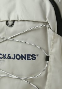Hellgrauer Rucksack aus robustem Gewebe mit schwarzen Akzenten. Verfügt über elastische Schnurdetaillierung und ein großes, dunkelblaues Logo.