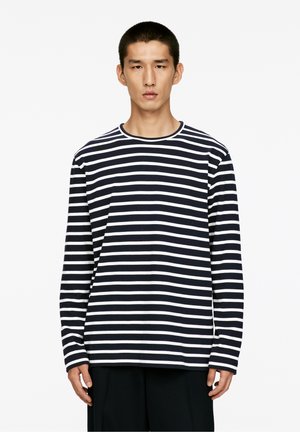 Junger Mann, der nach vorne blickt und ein schwarz-weiß gestreiftes langärmliges Hemd sowie schwarze Hosen trägt, vor einem schlichten weißen Hintergrund.