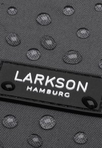 Ingrandimento di una borsa di stoffa nera con gocce d'acqua, dotata di un'etichetta rettangolare nera con la scritta "LARKSON HAMBURG" in caratteri bianchi. Superficie testurizzata.