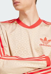 Bežs sporta krekls ar sarkaniem akcentiem, ar V-veida kakla izgriezumu, teksturētu rakstu un trim sarkanām svītrām gar pleciem. Uzrādīta Adidas emblēma.